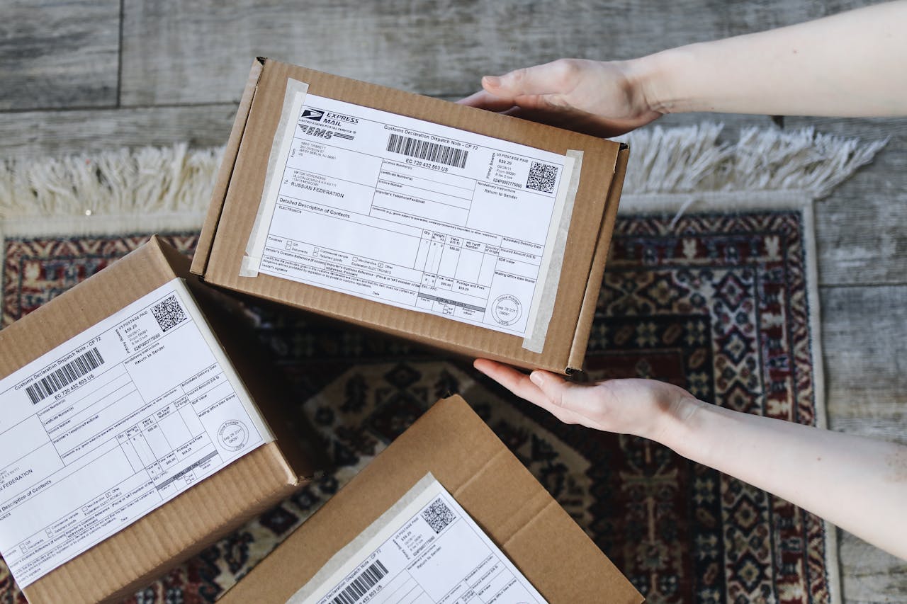 home-hero-1 Close-up of hands holding cardboard boxes with labels for delivery on a decorative carpet.